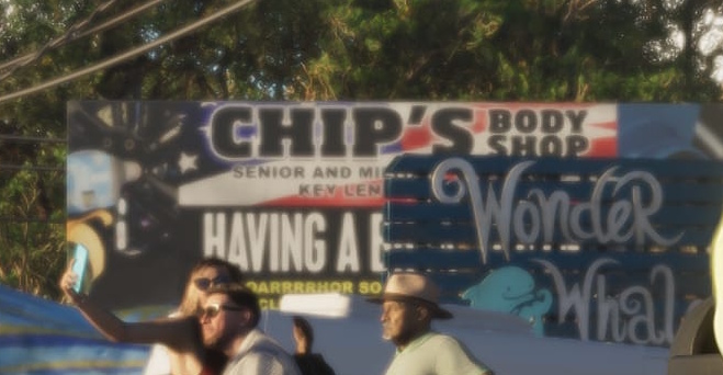 Люди на улице возле вывесок Chip's Body Shop и Wonder Whale, солнечный день, деревья на заднем плане, часть текста на английском видна на плакатах.