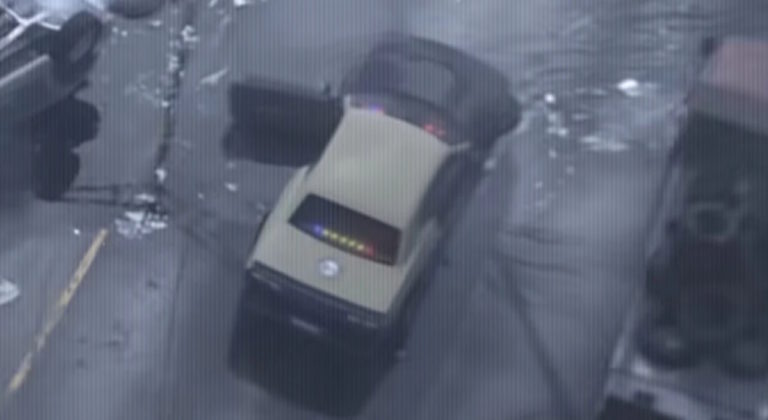 Полицейский автомобиль с включёнными мигалками стоит на мокрой дороге, водительская дверь открыта, вокруг видны следы мусора и другие машины. Ключевая фраза: полицейский автомобиль на дороге.
