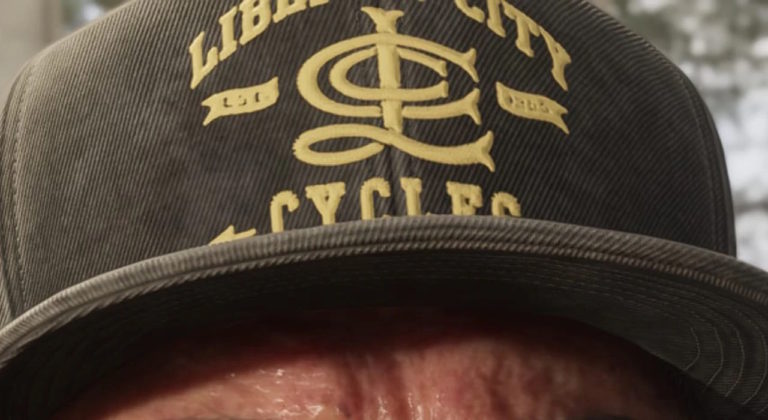 Крупный план кепки с надписью Liberty City Cycles и логотипом в центре, выполненной жёлтыми буквами на тёмной ткани. Видна часть лба человека под козырьком.