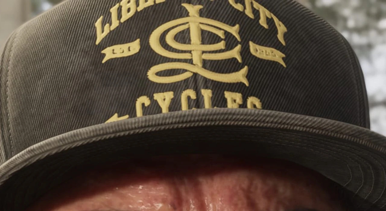Крупный план кепки с надписью Liberty City Cycles и логотипом в центре, выполненной жёлтыми буквами на тёмной ткани. Видна часть лба человека под козырьком.