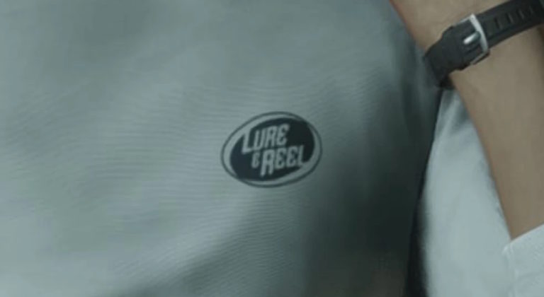 Светло-серая футболка с логотипом Lure & Reel в центре кадра, рядом видна часть руки с черными часами.