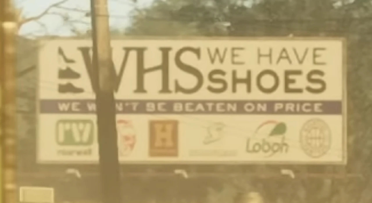 Рекламный щит с надписью WHS We Have Shoes, ниже слоган We Won't Be Beaten On Price и логотипы различных брендов. Щит расположен на улице, на фоне видны деревья и столб. Основная информация посвящена продаже обуви по выгодным ценам. Дизайн выполнен в светлых тонах, текст крупный и легко читается. Щит привлекает внимание прохожих и автомобилистов, информируя о большом ассортименте обуви.