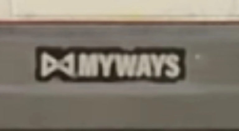 Надпись MYWAYS на черном фоне с белым логотипом слева, изображающим стилизованный бантик или галстук-бабочку. Надпись выполнена белыми буквами, фон вокруг надписи прямоугольный и выделяется на серой поверхности. Изображение слегка размытое, но текст хорошо различим. Возможно, это часть логотипа или брендовой наклейки, размещенной на транспортном средстве или оборудовании. Ключевая фраза: MYWAYS.