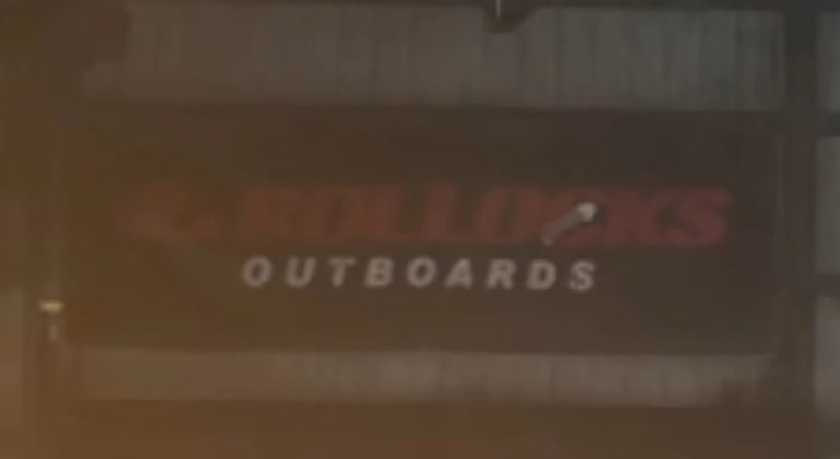 Вывеска с надписью "OUTBOARDS" и размытым красным текстом на тёмном фоне, часть которого неразборчива. Видна металлическая конструкция здания, освещение приглушённое, присутствует лёгкий блик в нижней части изображения. Атмосфера напоминает промышленную или складскую зону, где могут находиться мастерские или магазины, связанные с лодочными моторами. Ключевая фраза: вывеска магазина лодочных моторов.