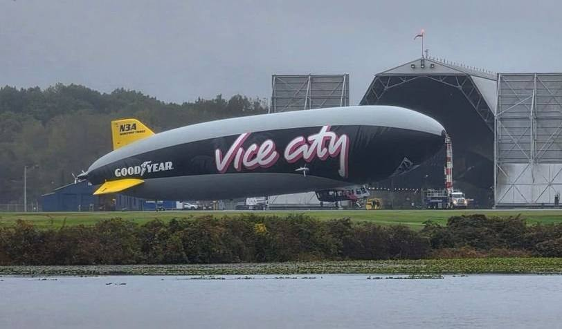 Дирижабль Goodyear с надписью Vice City стоит на аэродроме перед большим ангаром, на фоне деревья и пасмурное небо.